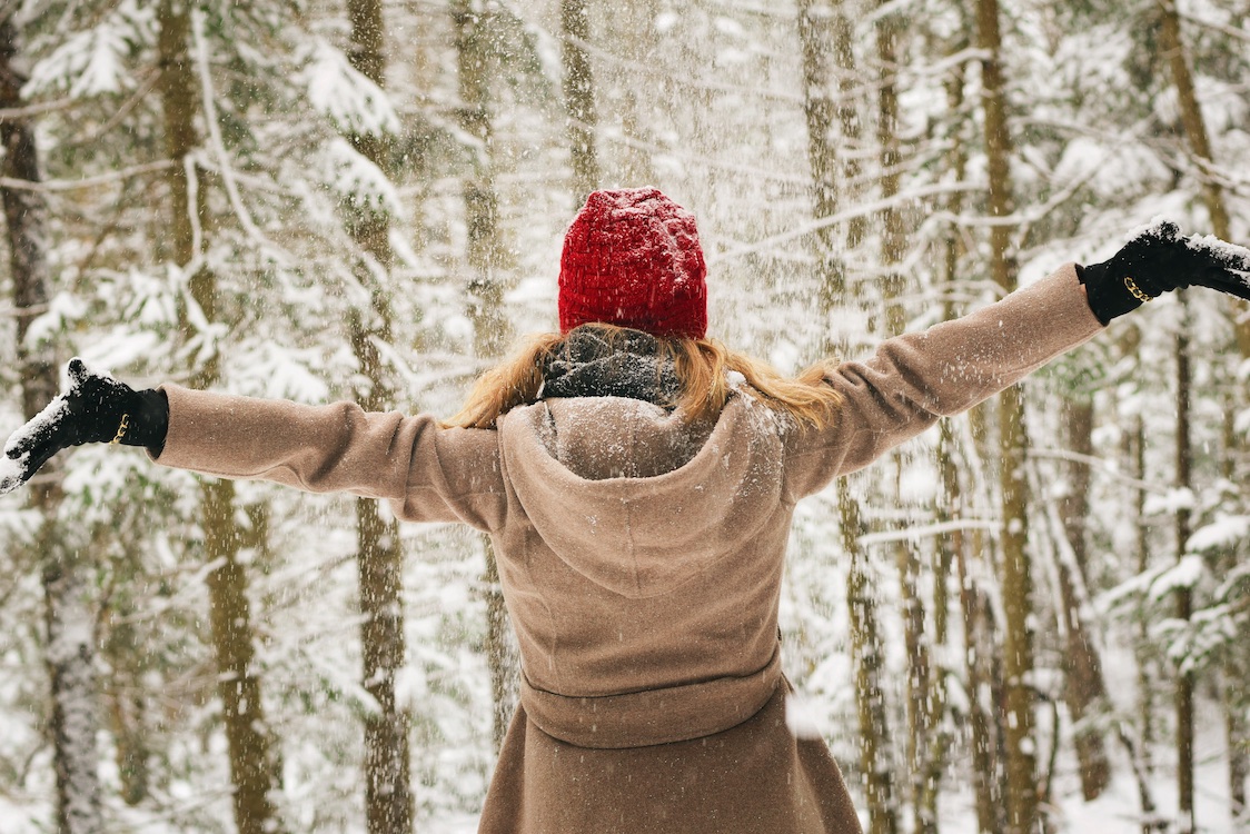 Courage Box Winter Program for Mental Wellness A woman in a jacket and red hat faces away from the camera with her arms in the air, facing a forest in winter.